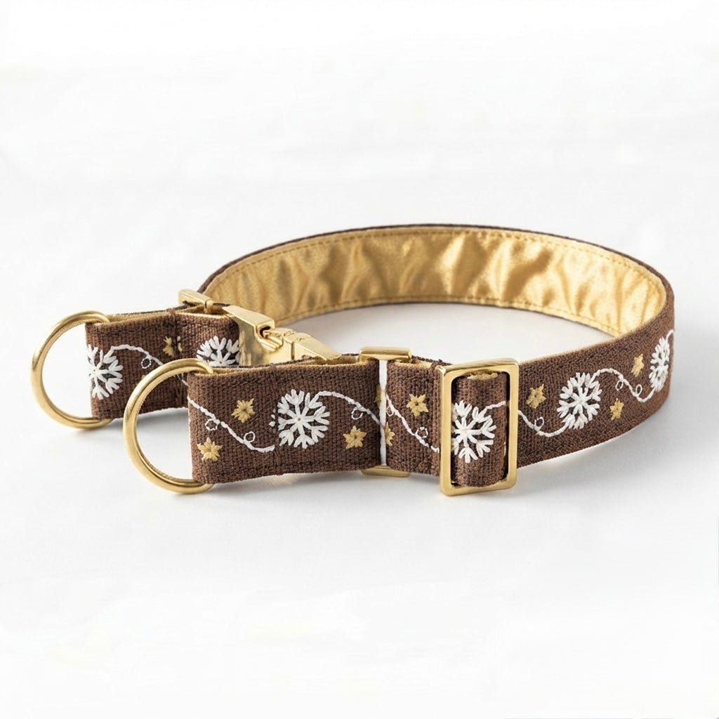 A brown dog collar with a gold satin lining, decorated with white snowflakes, gold stars, and white swirls. It features gold-toned hardware, including a D-ring, quick-release buckle, and clasp, shown on a clean white background.