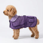 A small fluffy tan dog wearing a purple quilted pet coat with a purple-and-white gingham collar, photographed against a clean white studio background.