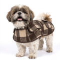 A small fluffy dog wearing a brown and beige plaid pet coat, standing against a solid white background.