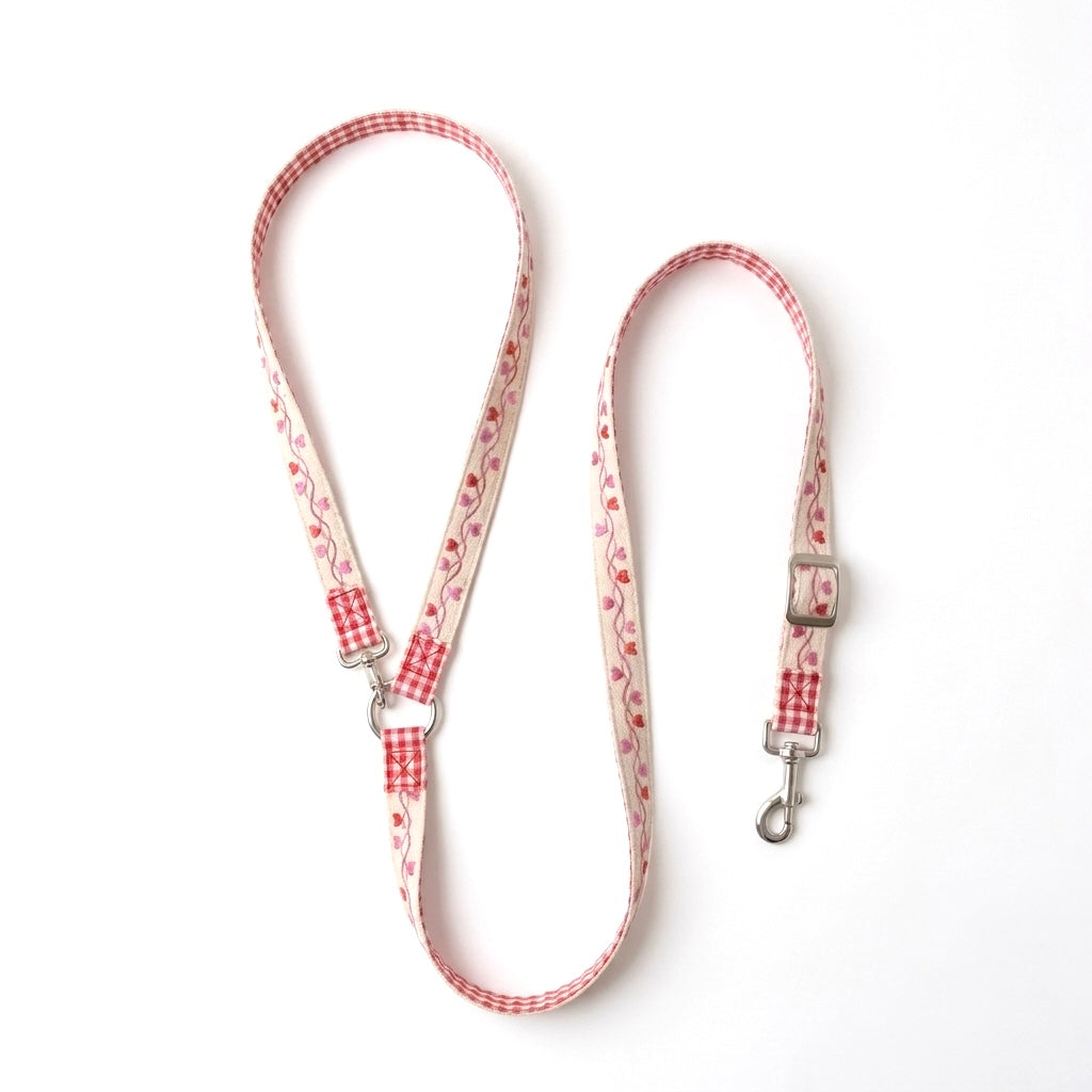 A fabric dog cross body leash with a red and white gingham pattern and a pink floral print, featuring silver metal hardware, laid out on a white background.