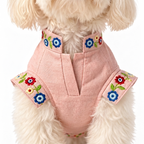 A small fluffy cream-colored dog wearing a light pink pet vest with colorful embroidered floral details on the collar and sides, set against a plain white background.