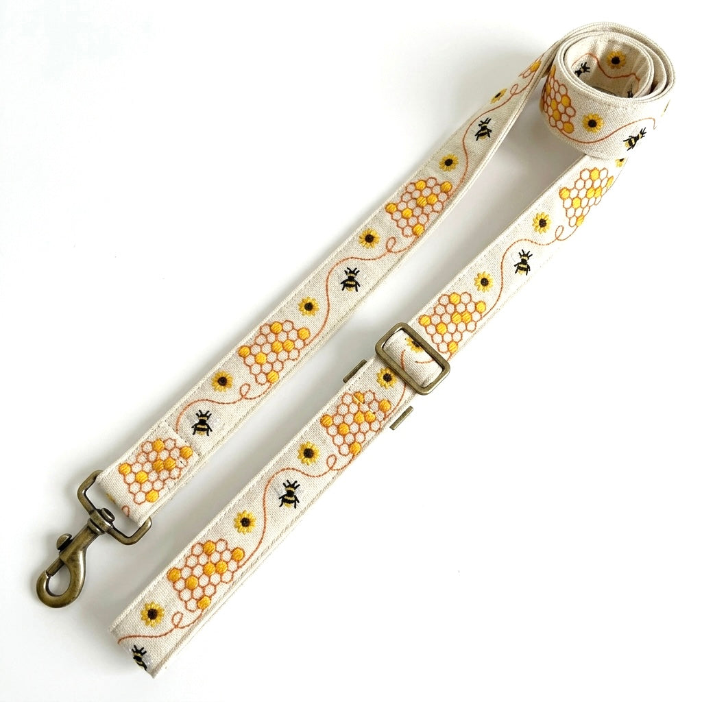 A fabric dog leash with a honeycomb and bee pattern on a cream background, featuring brass-colored metal hardware including a swivel clasp and a buckle.