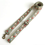 An adjustable fabric strap with a metal swivel clasp and buckle, featuring embroidered floral and berry patterns in red, light blue, and green on a muted sage-gray background.