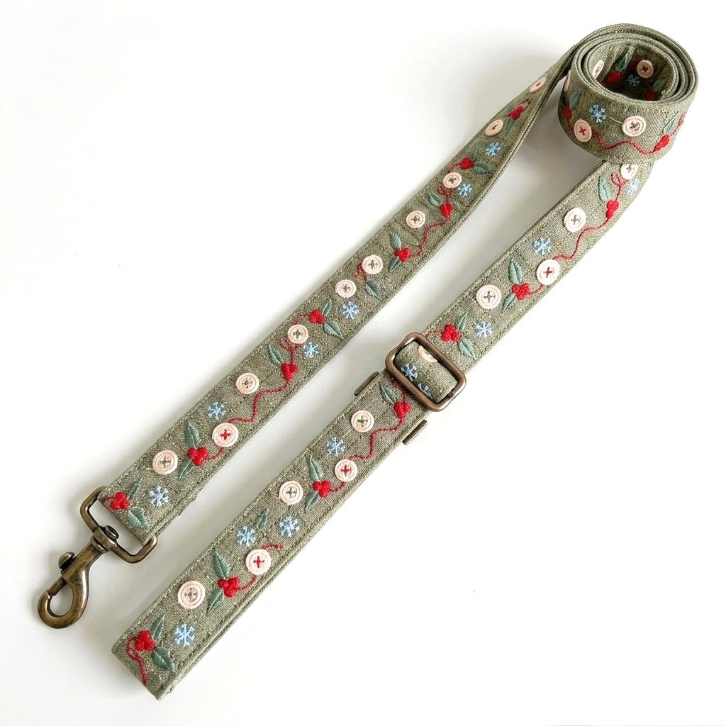 An adjustable fabric strap with a metal swivel clasp and buckle, featuring embroidered floral and berry patterns in red, light blue, and green on a muted sage-gray background.