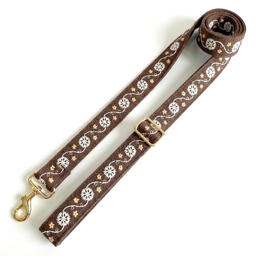 A brown leather dog lead strap with white floral and vine patterns and gold-tone hardware, including a snap hook and buckle, displayed on a clean white background.