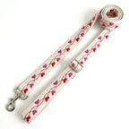 An off-white fabric dog leash with a repeating pattern of red and pink hearts on a wavy vine, featuring silver metal hardware including a buckle, snap hook, and adjustment slider, displayed on a clean white background.