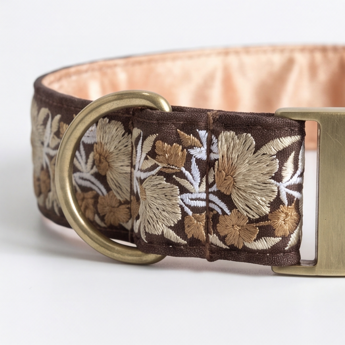 A close-up of a brown embroidered collar with a floral pattern in beige and white, featuring a brass ring and a matching brass buckle on a white background.