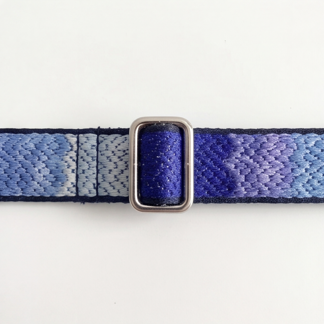 A close-up of a woven fabric strap with a blue and purple gradient pattern and a silver-toned metal buckle, shown against a plain white background.