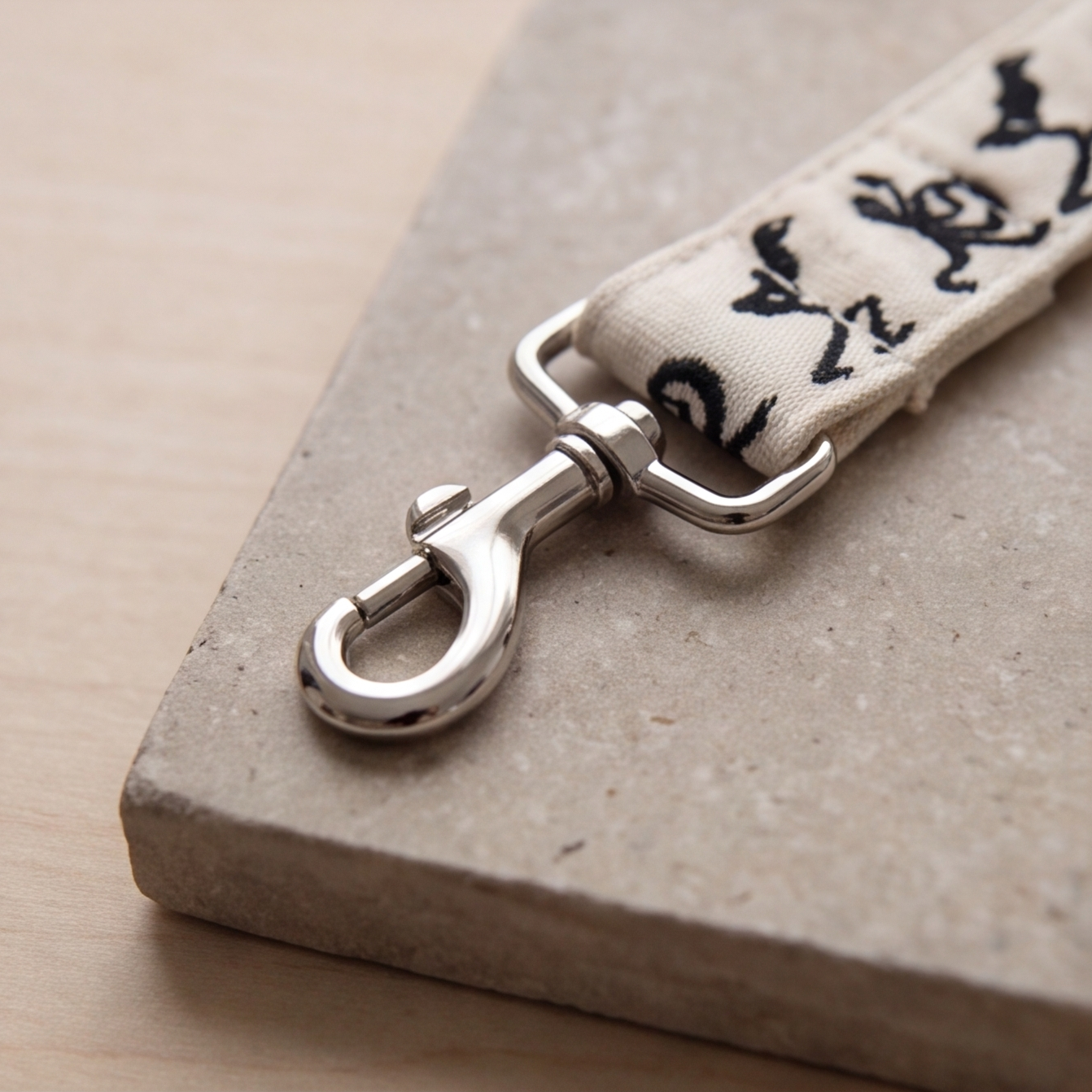 A close-up of a silver metal swivel clasp attached to a cream fabric strap with black printed script, resting on a textured stone tile over a light wood surface.