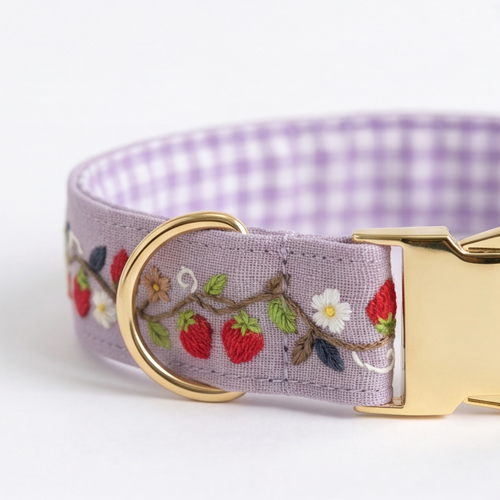 A close-up of a lavender pet collar featuring embroidered strawberries, white flowers, and green leaves on the front, with a purple and white gingham pattern lining and a gold-tone metal buckle.