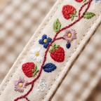 Close-up of a cream-colored woven strap featuring embroidered strawberries, blue and pink flowers, and green leaves on a red vine, set against a softly blurred beige background.