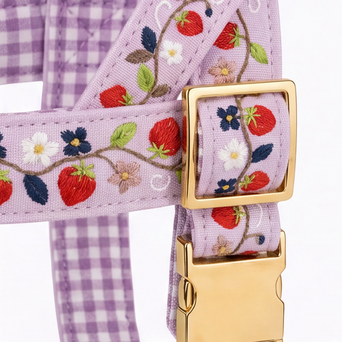 Close-up of a lavender pet harness with embroidered strawberries, flowers, and leaves on a light purple gingham fabric, featuring gold-tone metal hardware.