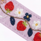 A close-up of a lavender embroidered fabric trim featuring red strawberries, white daisies, navy blue flowers, and dark blue leaves on a brown vine, set against a plain white background.