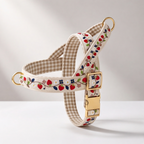 A cream-colored dog harness with a beige and white gingham check pattern, decorated with embroidered red strawberries, blue flowers, and green leaves. It features gold-tone metal hardware, including a buckle, rings, and a leash attachment point, shown on a clean white background.