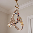 A fabric dog harness with a beige and white gingham pattern, decorated with a red cherry and green leaf print, featuring gold-tone hardware and a buckle, hanging from a gold hook.