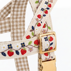 Close-up of a woven pet harness strap featuring embroidered strawberries, blue and white flowers, green leaves, and a beige-and-white gingham pattern, with a gold-tone metal buckle.