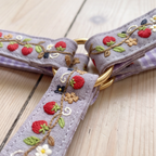 A close-up of a lavender-purple woven strap with embroidered red strawberries, green leaves, and gold-tone metal rings, laid on a light wood surface.