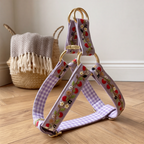 A dog harness with lavender and white gingham fabric, embroidered strawberries, and gold-tone metal rings, displayed on a wooden floor next to a woven basket.