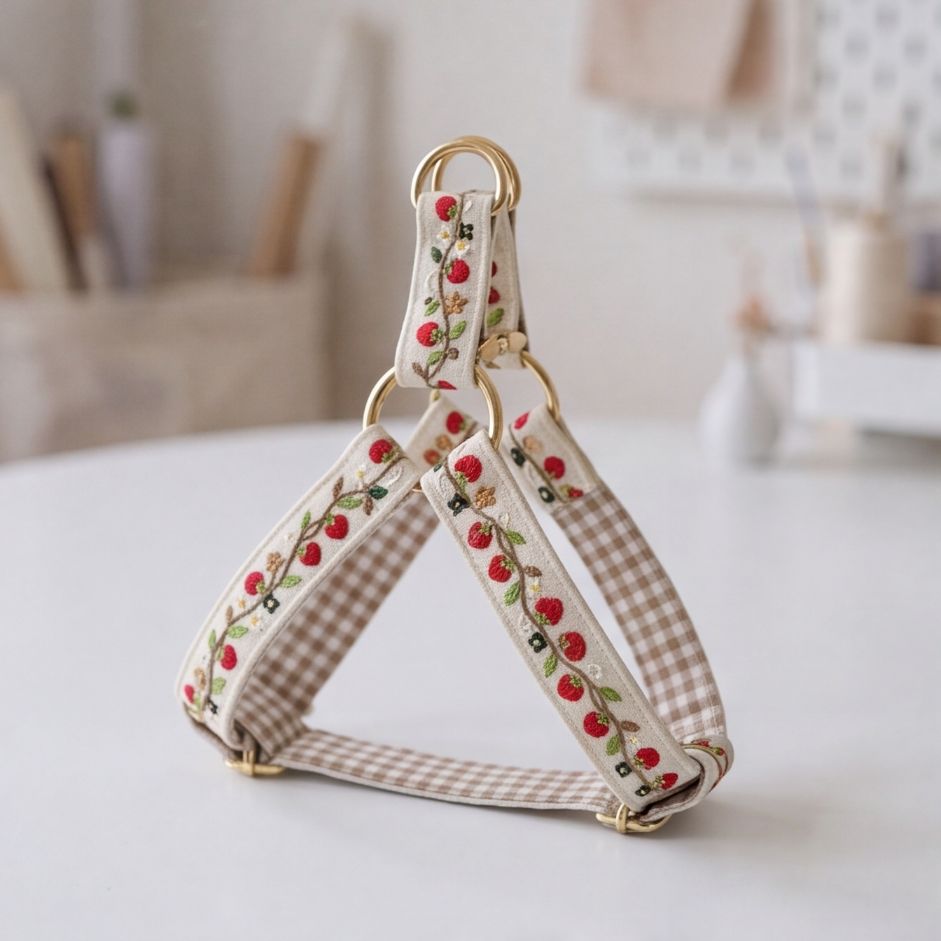 A dog harness with a cream and beige gingham pattern, decorated with embroidered red strawberries and green leaves, featuring gold-tone metal rings and adjusters, displayed on a white table with a soft-focus background.