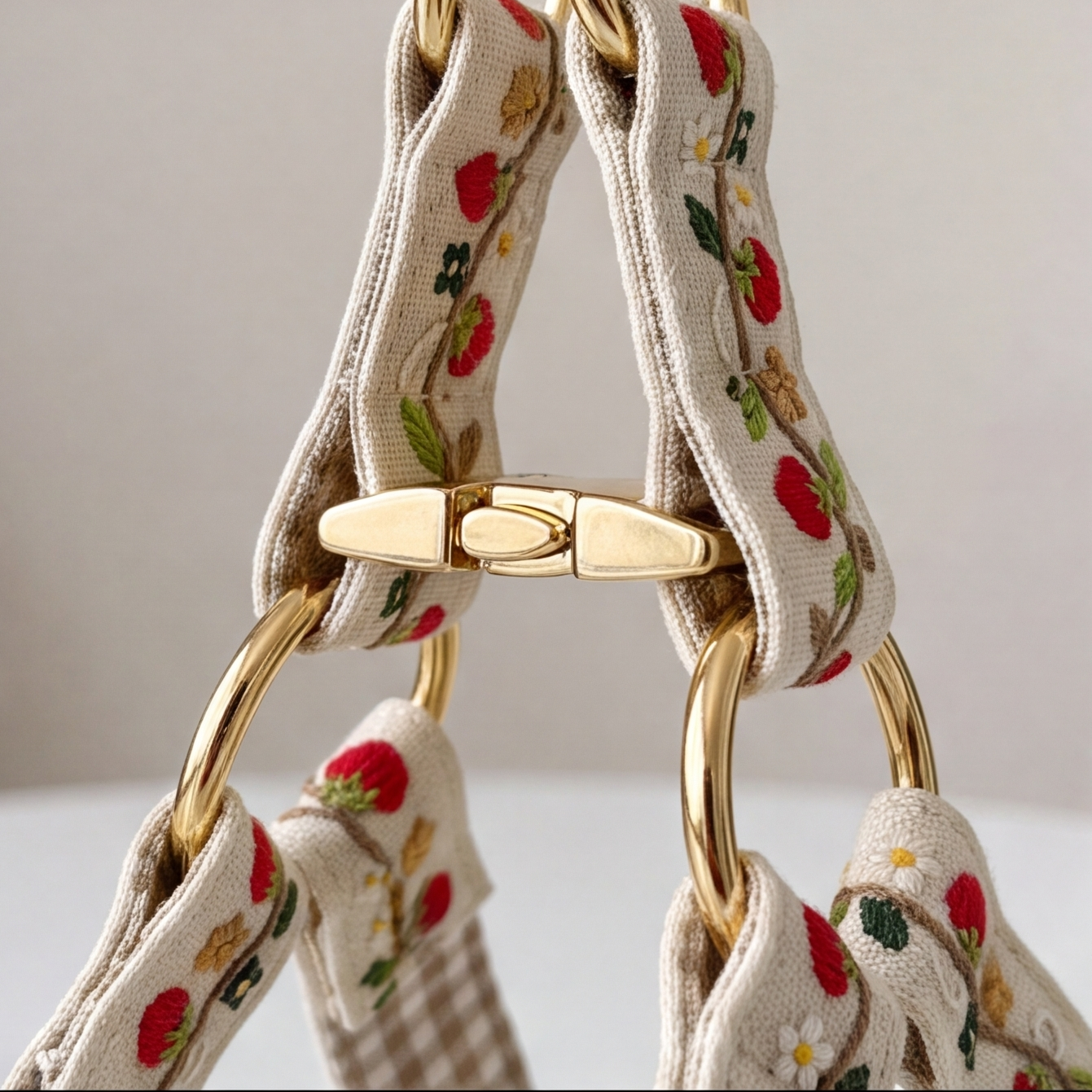 Close-up of a pet harness strap made of cream-colored fabric with a red strawberry and green leaf pattern, attached to gold-tone metal rings and a gold clasp.