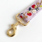A close-up of a lavender embroidered fabric strap with a shiny gold metal clasp, featuring floral and strawberry patterns.