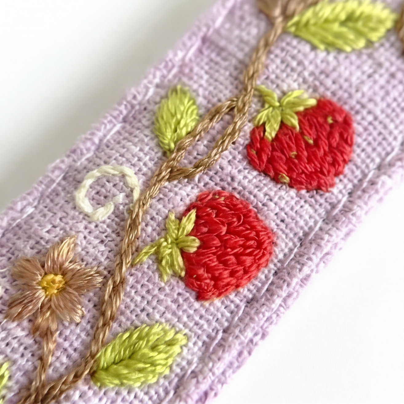 Close-up of a lavender embroidered fabric featuring red strawberries, green leaves, a gold vine, and a small white flower on a white background.