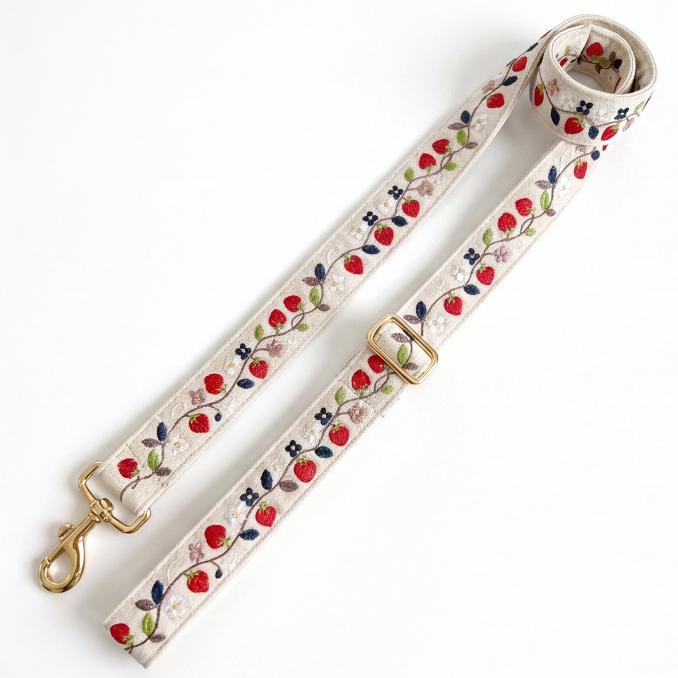 A woven leash strap with a floral and berry pattern, featuring a gold-tone swivel clasp and buckle, laid out on a white background.