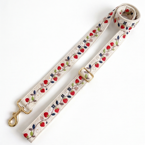 A woven leash strap with a floral and berry pattern, featuring a gold-tone swivel clasp and buckle, laid out on a white background.