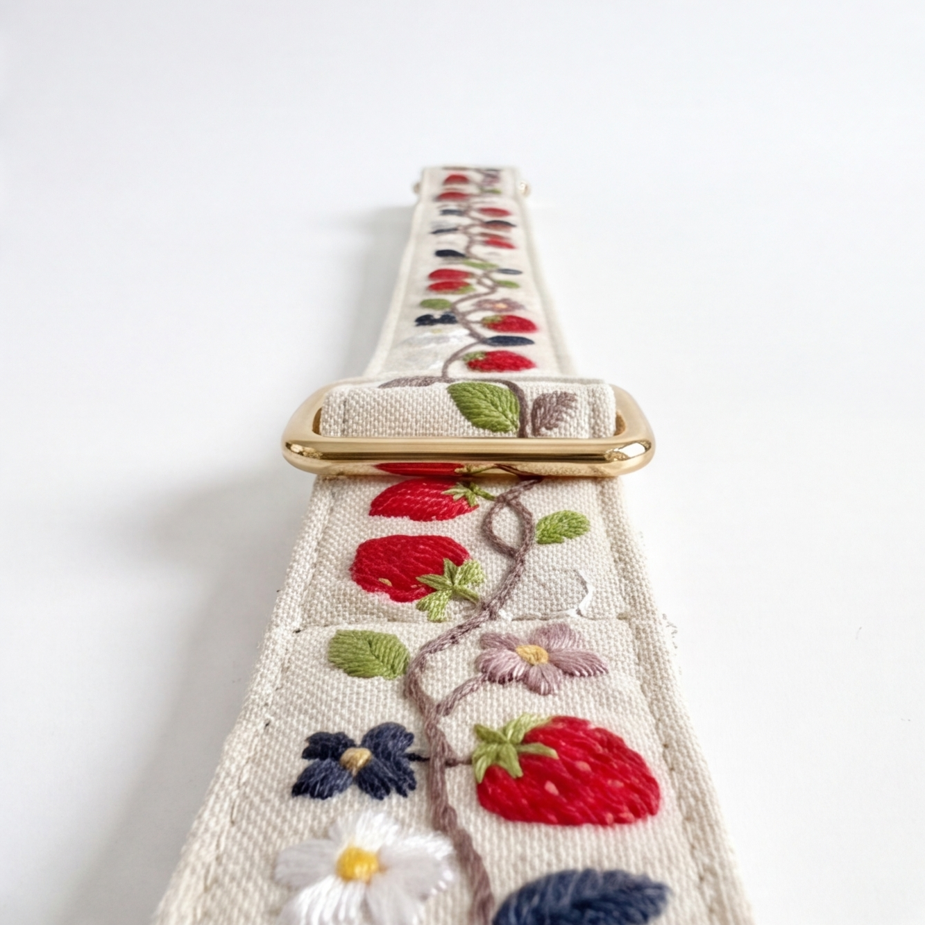 A close-up of a pet leash strap, likely a belt or bag strap, featuring embroidered strawberries, leaves, and flowers on a cream background with a gold metal buckle.