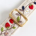 An embroidered fabric belt with a polished gold buckle, featuring red strawberries, green leaves, and white and purple flowers on a cream-colored woven background.