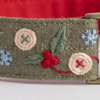 A close-up of a woven olive green fabric strap or belt featuring embroidered holiday motifs, including red berries, green leaves, beige floral rosettes, and light blue snowflakes, with a brass buckle partially visible on the right.