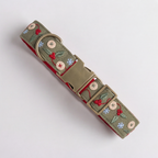 A dog collar with an olive green embroidered fabric featuring red berries, green leaves, and small blue and white flowers, complete with a brass buckle and D-ring, shown on a light gray background.