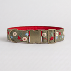 A dog collar with a sage green embroidered fabric exterior, featuring red berries, green leaves, and blue snowflake motifs, a bright red interior, and a brass-tone buckle.