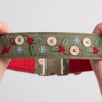 A close-up of a handmade olive green fabric belt being held by two hands. The belt is decorated with embroidered green leaves, red berries, blue snowflakes, and beige wooden buttons.