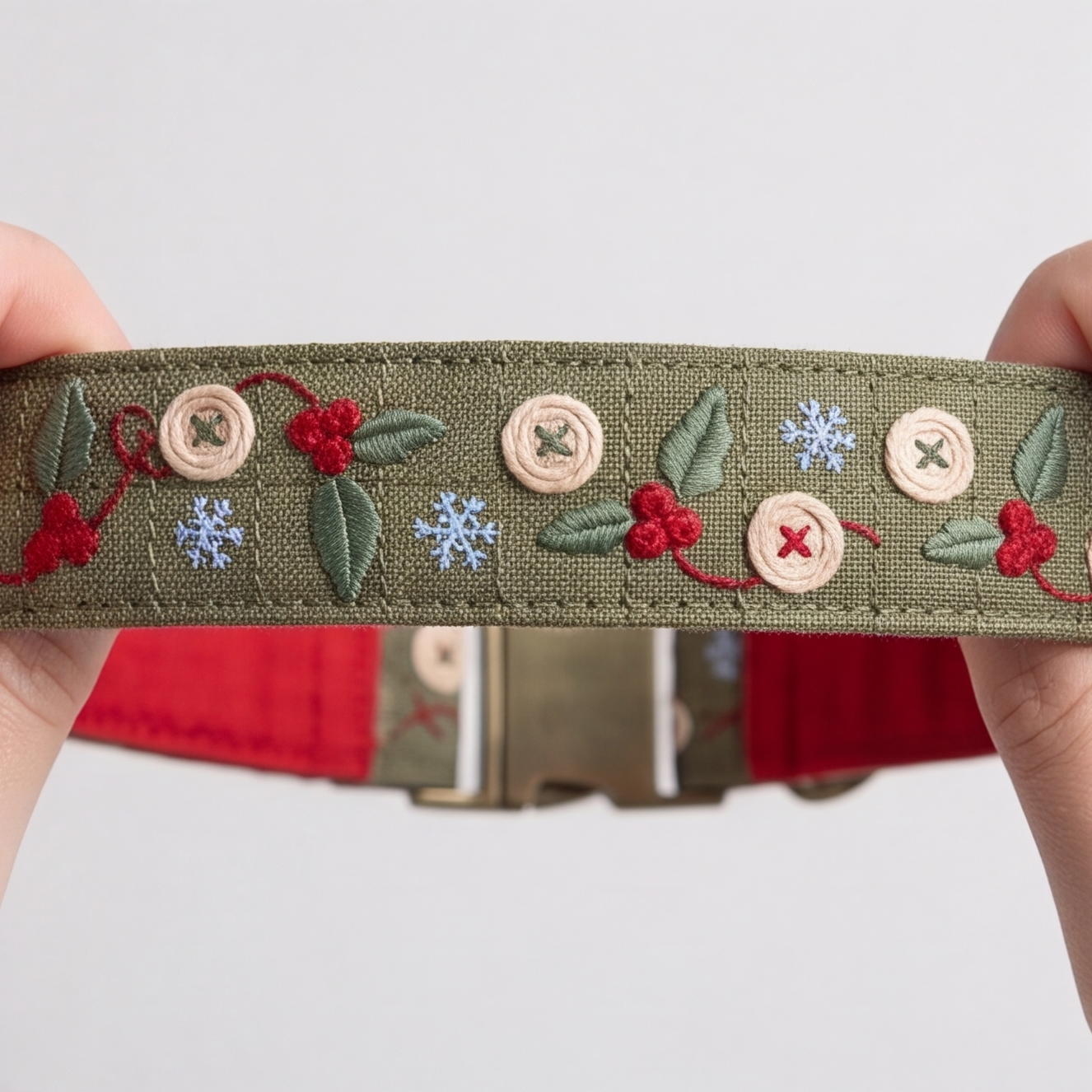 A close-up of a handmade olive green fabric belt being held by two hands. The belt is decorated with embroidered green leaves, red berries, blue snowflakes, and beige wooden buttons.