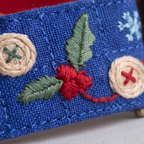Close-up of a blue embroidered fabric item featuring a red berry motif with green leaves, cream buttons with red and green crosses, and a light blue snowflake detail.