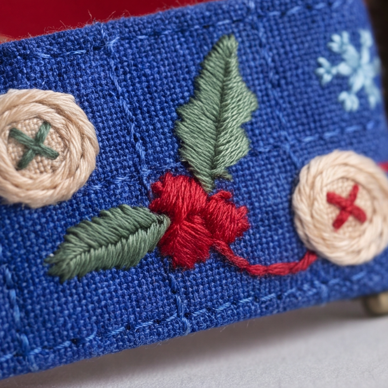 Close-up of a blue embroidered fabric item featuring a red berry motif with green leaves, cream buttons with red and green crosses, and a light blue snowflake detail.