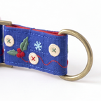 A close-up of a blue fabric strap with embroidered holiday motifs, including holly leaves, red berries, a snowflake, and decorative buttons, attached to a brass ring.