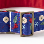 A close-up of a blue fabric pet collar with an antique bronze buckle, decorated with embroidered red berries, green leaves, white snowflakes, and beige buttons.