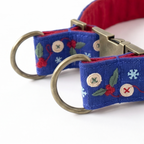 A close-up of a blue fabric pet collar with embroidered red berries, green leaves, snowflakes, and button motifs, featuring antique brass hardware and a red interior lining.