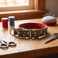 A fabric dog collar with a green floral pattern and red lining, displayed on a wooden table surrounded by sewing supplies like spools of thread, scissors, and a pin cushion.