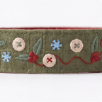 An embroidered olive green fabric trim or ribbon featuring decorative motifs including pink buttons, red berries, blue snowflakes, and green leaves on a white background.