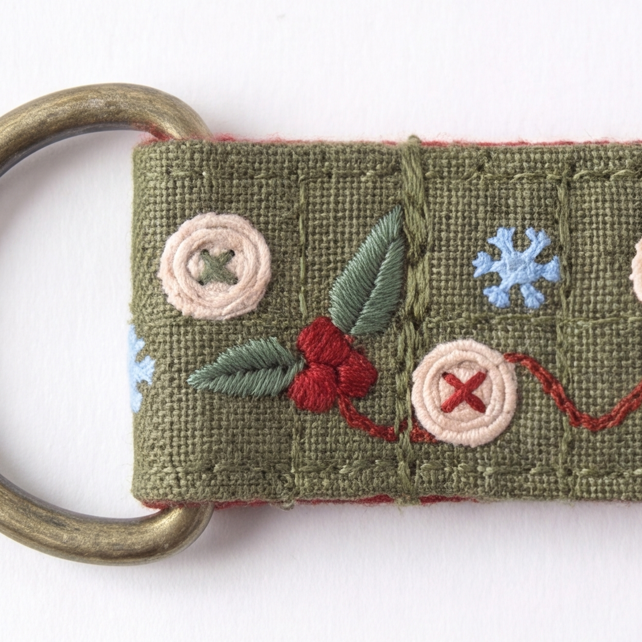 An embroidered olive green fabric key fob with a brass ring, featuring festive Christmas motifs including red berries, green leaves, beige buttons, and a light blue snowflake on a white background.