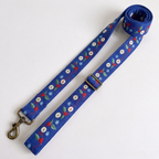 A blue fabric dog leash with a repeating pattern of red berries, green leaves, white snowflakes, and white paw prints, featuring brass-colored metal hardware including a swivel clasp and adjustment buckle, shown on a plain white background.