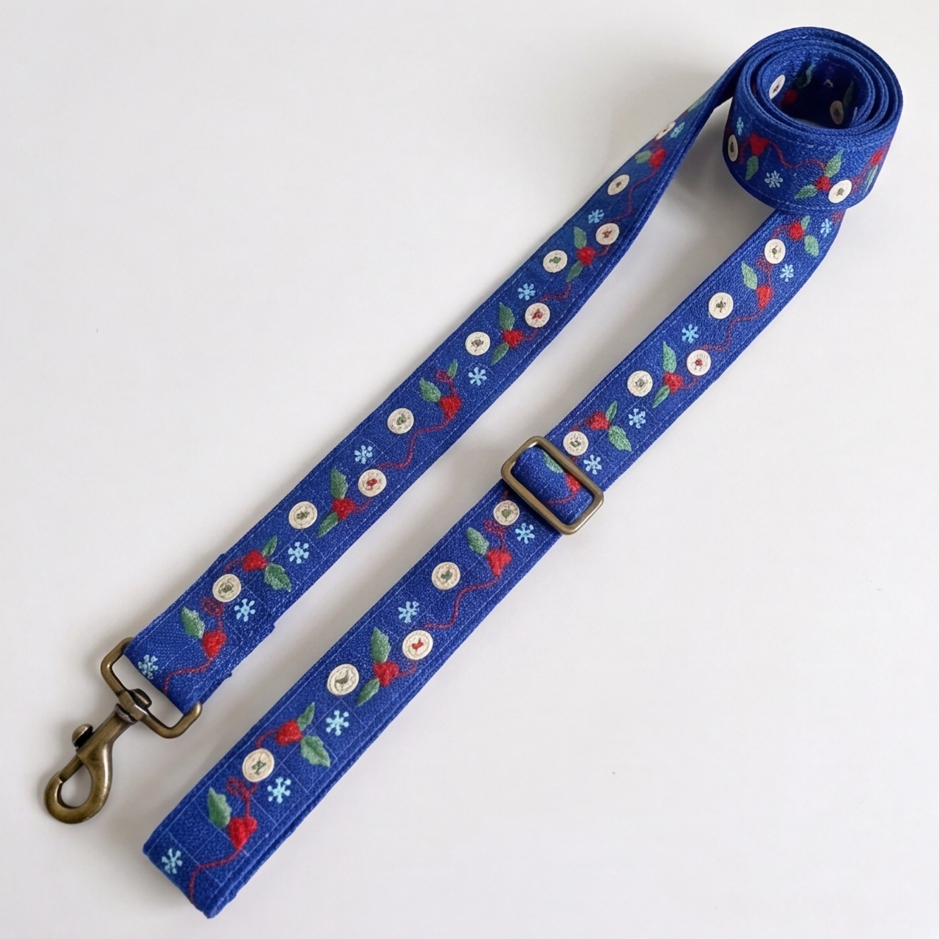 A blue fabric dog leash with a repeating pattern of red berries, green leaves, white snowflakes, and white paw prints, featuring brass-colored metal hardware including a swivel clasp and adjustment buckle, shown on a plain white background.