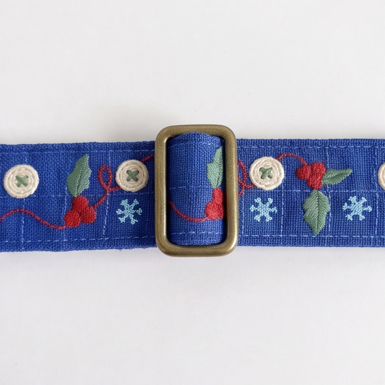 A close-up of a blue woven belt with embroidered winter motifs, including red berries, green leaves, light blue snowflakes, and white decorative buttons, featuring a central brass buckle.