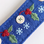 A close-up of a blue woven fabric strap or belt featuring embroidered holiday motifs, including green leaves, red berries, light blue snowflakes, and a cream-colored decorative button.