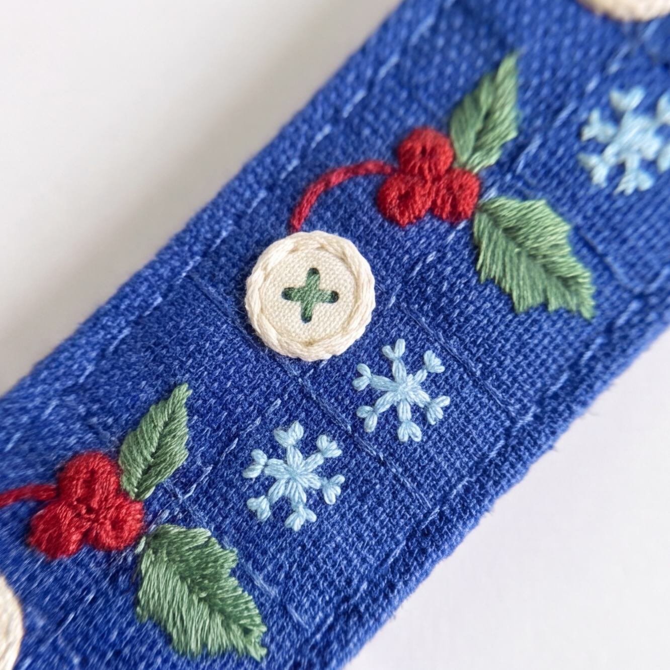 A close-up of a blue woven fabric strap or belt featuring embroidered holiday motifs, including green leaves, red berries, light blue snowflakes, and a cream-colored decorative button.