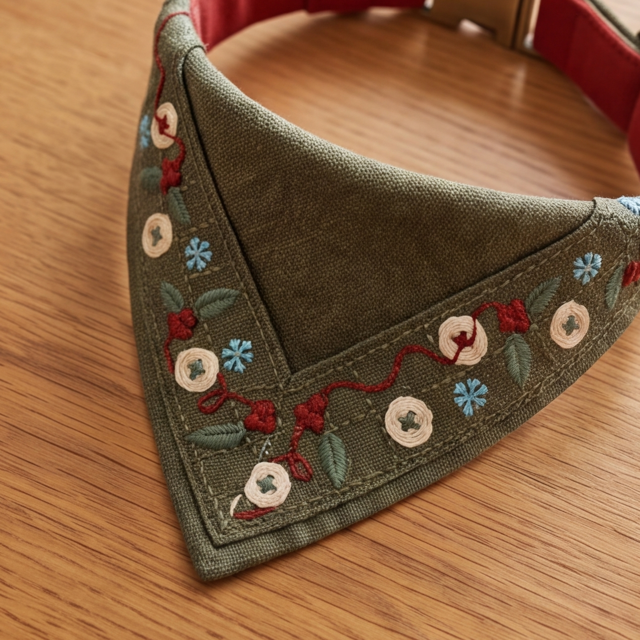 An olive green triangular bandana collar with floral embroidery, displayed on a wooden surface.