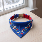 A blue pet bandana collar with red lining and embroidered floral details, displayed on a light wood table near a window.